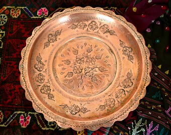 Könnte beinhalten: Ein großes, rundes Serviertablett aus Kupfer mit aufwendigen floralen und geometrischen Mustern, die in die Oberfläche eingeätzt sind. Das Tablett hat einen gewellten Rand und liegt auf einem bunten, gemusterten Stoff.