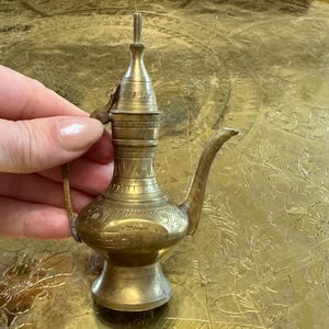 May include: A small, metallic brass-colored oil lamp or vessel featuring an elongated spout, a narrow neck, and a flared base. The surface of the object is decorated with intricate, hand-etched linear patterns and geometric engravings throughout.