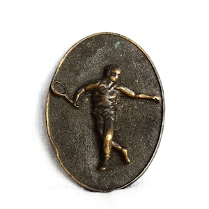 Peut inclure: Une boucle de ceinture vintage en bronze avec une image en relief d'une personne jouant au tennis. La personne tient une raquette de tennis et est en plein swing.