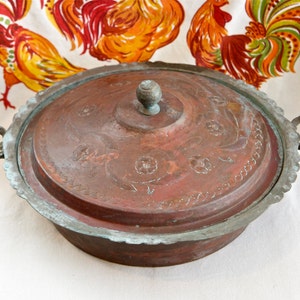 May include: An antique copper serving dish with a lid. The dish features a decorative floral design and two handles. The lid has a knob on top.
