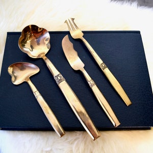 May include: Set of four gold-toned metal serving utensils with ornate details. The set includes a ladle, a small spoon, a butter knife, and a fork.