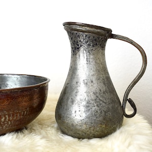 May include: A silver pitcher with an intricate engraved design and a copper bowl with a similar design. Both are on a white fluffy surface.