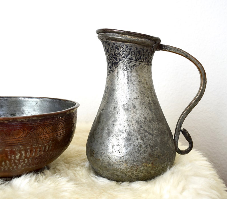 May include: A silver pitcher with an intricate engraved design and a copper bowl with a similar design. Both are on a white fluffy surface.