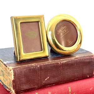 May include: Two polished brass picture frames, one rectangular and one circular. The circular frame features text reading Solid Brass 2 1/2 x 2 1/2. Both frames have a smooth, reflective metallic finish and are designed to hold small photographs.