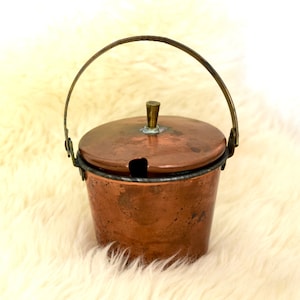 May include: A small copper sugar bowl with a lid and a handle. The bowl is sitting on a white fluffy surface.