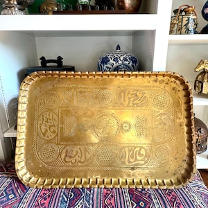 May include: A rectangular brass tray with a scalloped edge and a central circular medallion. The surface features intricate hand-engraved patterns, including geometric textures and stylized Arabic calligraphy arranged in a symmetrical border.