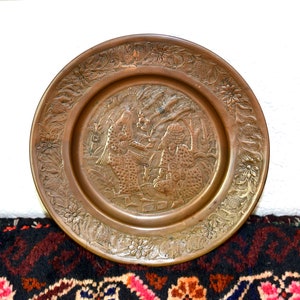 May include: A round, brass plate with an intricate design featuring two figures in a garden setting. The plate has a raised, embossed design and a decorative border.