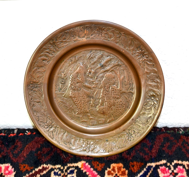 May include: A round, brass plate with an intricate design featuring two figures in a garden setting. The plate has a raised, embossed design and a decorative border.