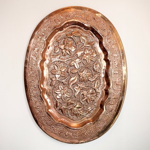 May include: Oval copper tray with intricate floral and leaf patterns. The tray has a raised, detailed design with a polished finish. The border features a delicate, repeating pattern. The tray is a decorative piece.