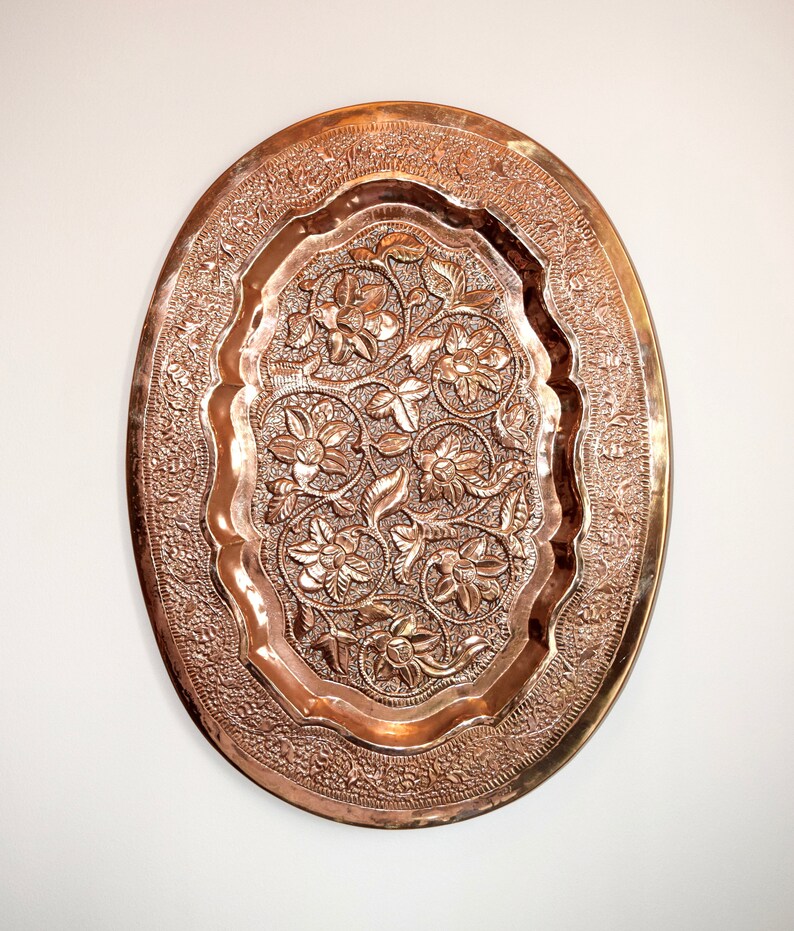 May include: Oval copper tray with intricate floral and leaf patterns. The tray has a raised, detailed design with a polished finish. The border features a delicate, repeating pattern. The tray is a decorative piece.