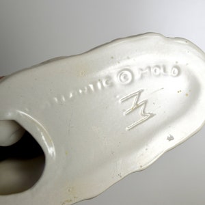 May include: White ceramic figurine with a brown base. The bottom of the figurine is marked "Plastic &copy; Holo" and a stylized "Z" logo.