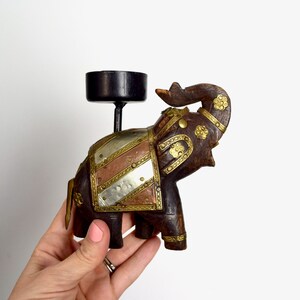 May include: A wooden elephant candle holder with metal accents. The elephant is dark brown with gold and silver accents. The candle holder is a black metal cup on top of the elephant's head.