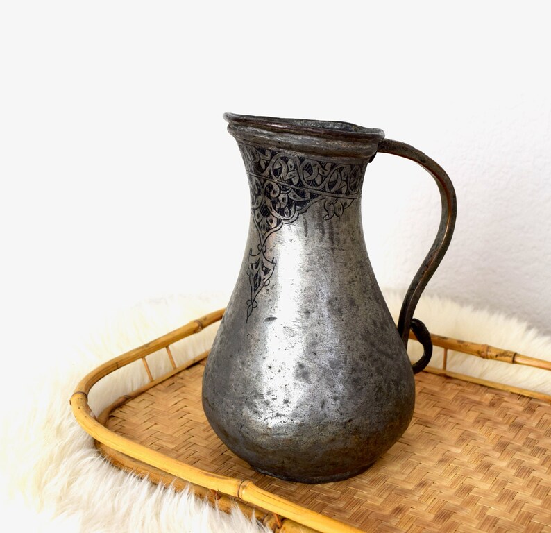 May include: A silver pitcher with an intricate black design on the top. The pitcher is sitting on a woven bamboo tray.