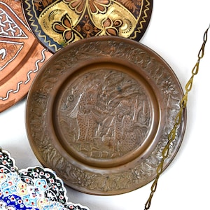 May include: A decorative copper plate with an intricate design of flowers and figures. The plate is hanging from a brass chain.