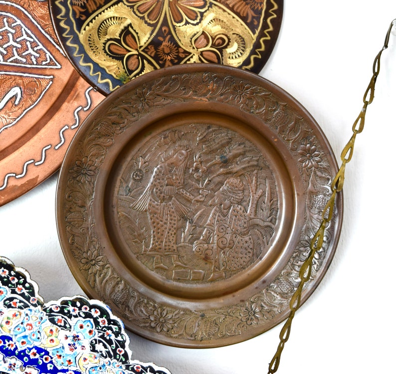 May include: A decorative copper plate with an intricate design of flowers and figures. The plate is hanging from a brass chain.