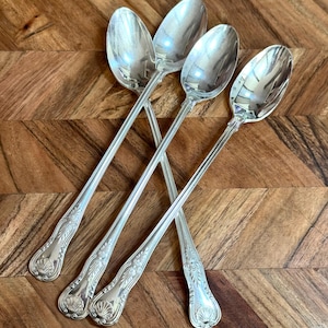May include: Four silver-toned long-handled spoons with polished oval bowls and slender stems. The handles feature a decorative embossed shell motif at the base and a vertical grooved pattern extending up the length of the handle toward the bowl.
