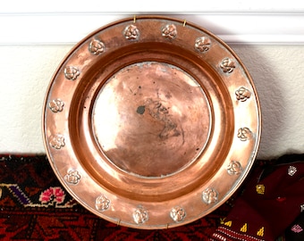May include: A large, round copper serving dish with a hammered finish and a raised rim. The dish is decorated with a repeating pattern of embossed floral designs.