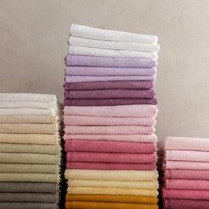 Könnte beinhalten: Drei Stapel gefalteter rechteckiger Stoffservietten in einem Farbverlauf. Der linke Stapel zeigt Weiß, Creme, Beige, Oliv und Grau. Der mittlere Stapel umfasst Weiß, Lavendel, Violett, Rosa, Rot und Gelb. Der rechte Stapel zeigt Rosa und Rot.