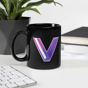 May include: A glossy black ceramic mug featuring a large, stylized letter V on the front. The letter V is designed with a gradient effect, transitioning from white to light pink and deep purple, creating a three-dimensional appearance on the mug.