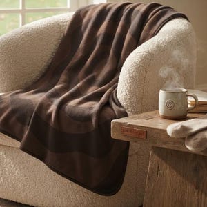 Puede incluir: Una manta suave de color marrón oscuro con un patrón ondulado tonal drapeada sobre un sillón texturizado de color crema. La manta presenta una textura suave similar al vellón y está colocada junto a una mesa auxiliar de madera.