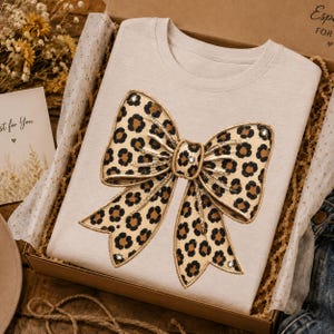 Pode incluir: Uma t-shirt de gola redonda em bege claro com um gráfico grande e centralizado de um laço com padrão de leopardo castanho e preto. O design do laço inclui pequenos strass transparentes e brilhantes no centro e nos quatro cantos.