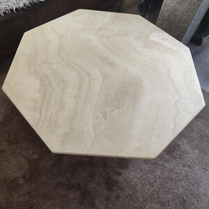 Op de afbeelding: Een achthoekige salontafel met een glad blad van lichtbeige travertijnsteen, voorzien van natuurlijke aders in crème- en bruintinten. Het oppervlak vertoont een kenmerkende gelaagde textuur die typerend is voor natuursteen.