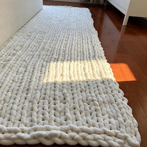 May include: A rectangular, off-white chunky knit runner rug featuring a thick, hand-knitted texture with large, interlocking loops. The rug is crafted from soft, plush yarn and displays a consistent, uniform pattern throughout its length.