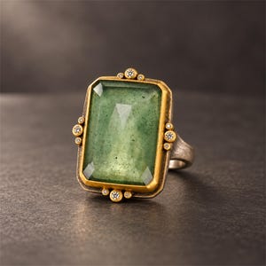 May include: A rectangular ring featuring a large, faceted green stone set in a gold-toned bezel. The setting is accented with small, clear round stones arranged in clusters along the top, bottom, and sides of the central green gemstone.
