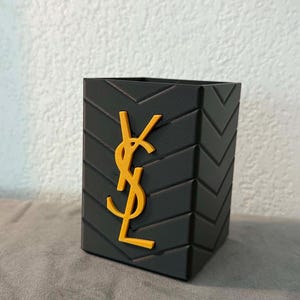 Peut inclure: Un organisateur de bureau noir de forme carrée présentant un motif à chevrons en relief sur ses surfaces extérieures. Le panneau avant affiche un emblème jaune YSL proéminent, orienté verticalement, centré sur la surface noire texturée.