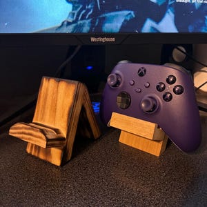 May include: A purple wireless video game controller resting on a light-colored wooden stand next to an empty, scorched-finish wooden stand. Both stands feature an A-frame design with a horizontal ledge for holding devices or controllers.