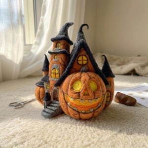 Op de afbeelding: Een naaldgevilte Halloween-decoratie bestaande uit een oranje kasteelstructuur met zwarte puntdaken en een grote, lachende pompoen aan de voorzijde. Het object heeft gele ramen en grijze treden en is vervaardigd uit zachte wolvezels.