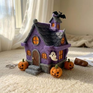 May include: A needle-felted purple haunted house with a dark gray roof, orange windows, and a black bat on top. A small white ghost hangs from the side, and three orange jack-o'-lanterns with carved faces sit at the base of the structure.
