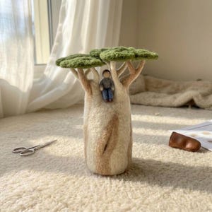 May include: A needle-felted sculpture of a baobab tree with a thick, light beige trunk and a wide, textured green canopy. A small, grey-clad felt figure sits nestled between the branches of the tree. The sculpture has a soft, woolly appearance.
