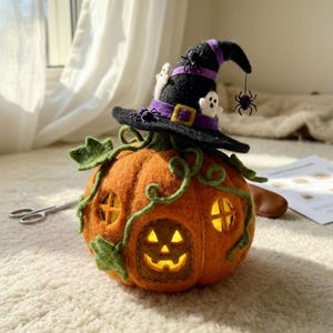 May include: A needle-felted orange pumpkin house with a glowing interior, featuring green felt vines and a black witch hat topper. The hat is decorated with a purple band, a yellow buckle, two small white ghosts, and two black spider accents.