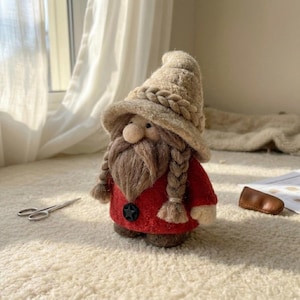 May include: A needle-felted gnome figurine featuring a beige pointed hat with a braided trim, a long brown beard with two side braids, and a red tunic adorned with a single black star-shaped button. The gnome has a round nose and brown feet.
