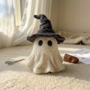 Puede incluir: Una figura de fantasma de lana fieltrada en color blanco roto, con dos ojos circulares oscuros y un sombrero de bruja puntiagudo y texturizado en gris carbón. El fantasma tiene un aspecto suave y drapeado con pliegues en la base, creando una forma redondeada.