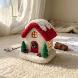 May include: A needle-felted house ornament featuring a cream-colored base, a bright red roof covered in white snow, and a red front door with a green wreath. Two green felted pine trees with red berries flank the door, and warm light glows from windows.