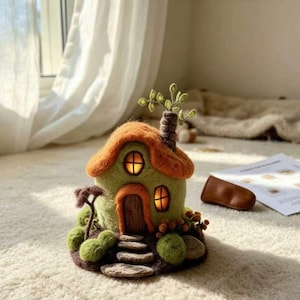 May include: A needle-felted house sculpture featuring a light green structure with an orange roof, a brown door, and two illuminated windows. The base includes felted green mounds, stone-like steps, and a small tree with thin branches.