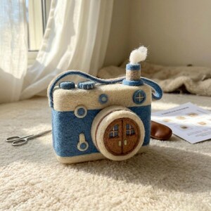 May include: A needle-felted camera-shaped object in cream and blue wool. The lens is a brown circular door with a small chimney on top emitting a white puff of wool. It features blue circular accents, a strap, and a textured, soft-touch finish.