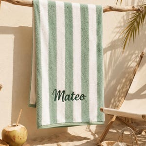 Puede incluir: Una toalla de playa rectangular con rayas verticales en color verde salvia y blanco. La toalla está bordada con el nombre Mateo en una fuente cursiva de color verde oscuro. El tejido tiene una superficie suave y texturizada de algodón.