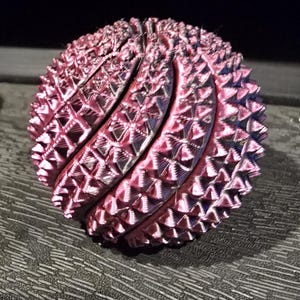 May include: A spherical object with a metallic pink finish and a textured, spiky surface. The sphere is composed of multiple interlocking segments that create a repeating geometric pattern across its entire rounded exterior surface.