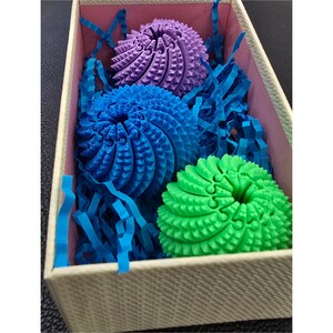 May include: Three spherical, textured objects in purple, blue, and green are nestled inside a rectangular box filled with blue paper shred. Each sphere features a complex, interlocking geometric pattern with small, pointed protrusions across its surface.