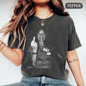 May include: A charcoal gray short-sleeve t-shirt featuring a black and white graphic print of a person giving the middle finger. The shirt has a crew neckline and a vintage-style washed finish. The text PEPPER is printed in the top right corner.