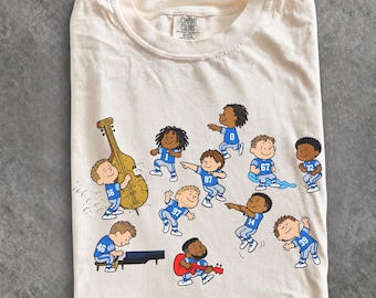 May include: An ivory Comfort Colors t-shirt featuring a graphic of cartoon characters wearing blue football jerseys with numbers, playing musical instruments including a double bass, piano, and red guitar, with the text COMFORT COLORS IVORY.