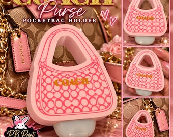 May include: A pink silicone pocketbac holder shaped like a handbag with a repeating circular pattern and the word COACH printed in gold. The holder features a cutout handle design and a white base, designed to hold a small hand sanitiser bottle.