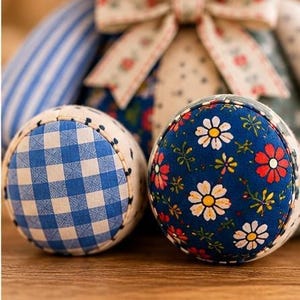 May include: Two round fabric pincushions featuring distinct patterns. One displays a blue and white gingham check, while the other showcases a dark blue background with a floral print of white daisies and red flowers. A fabric bow sits above.