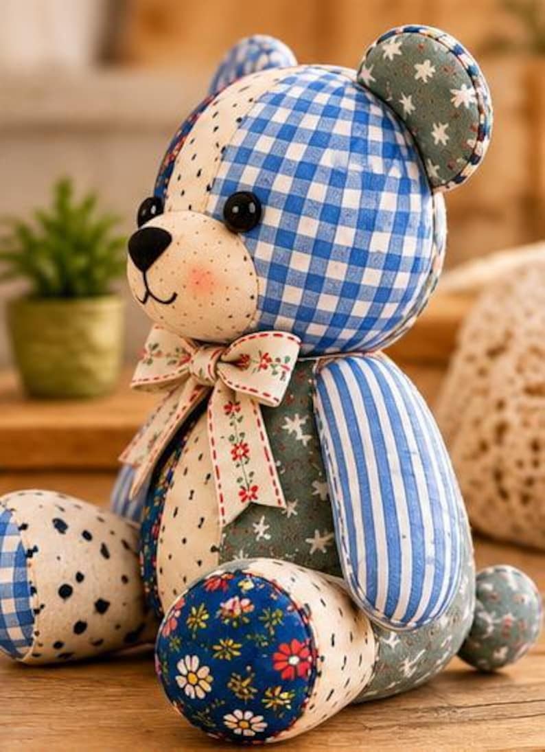 May include: A patchwork teddy bear crafted from various fabric patterns including blue and white gingham, blue stripes, floral prints, and star motifs. The bear features black button eyes, a black stitched nose, and a cream-coloured bow with red flowers.
