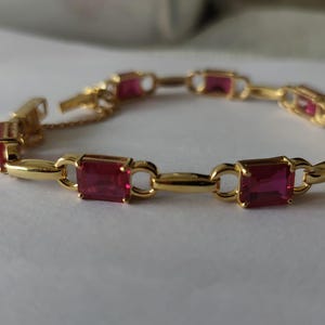 May include: A gold-toned link bracelet featuring alternating rectangular red gemstones and smooth gold-toned metal bars. The bracelet is constructed with a series of connected segments and includes a secure clasp mechanism with a small safety chain attached.