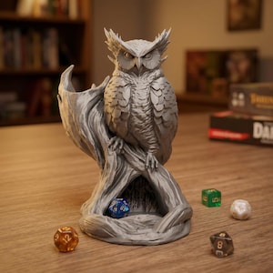 May include: A silver-colored 3D printed owl figurine perched on a textured tree stump base. The base features a hollow cavity containing a blue multi-sided die. Three additional multi-sided dice in orange, green, and white are placed nearby.