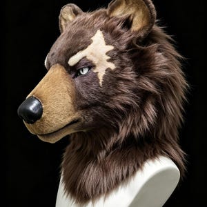 Könnte beinhalten: Eine Bärenmaske aus braunem Kunstfell mit beigefarbener Schnauze, schwarzer Nase und einer gezackten cremefarbenen Markierung über dem Auge. Die Maske hat spitze Ohren, strukturiertes Fell und ein weißes Auge mit Pupille auf einer weißen Büste.